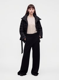 Femme debout portant une chemise beige boutonnée, une doudoune noire, un pantalon large noir, les mains dans les poches sur un fond uni.
