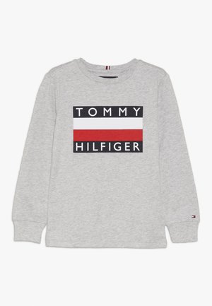 Jasnoszara koszula z długim rękawem z napisem „Tommy Hilfiger” i prostokątnym logo w kolorach czerwonym, białym i czarnym, umieszczonym na środku klatki piersiowej.
