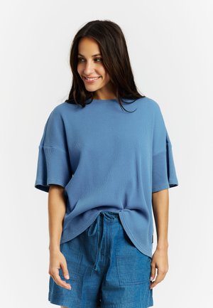 Jonge vrouw met lang, donker haar draagt een losse blauwe top met korte mouwen en denim short met trekkoord, lacht en kijkt naar links.