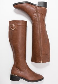 Bottes hautes jusqu'au genou en similicuir marron, à bout rond et talon bloc bas, équipées d'une fermeture éclair latérale et d'une sangle décorative avec un anneau en métal.