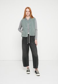 Colete verde de lã com botões frontais, camisa cinza de manga comprida por baixo, combinado com calças largas pretas e ténis pretos, em pé sobre um fundo branco.