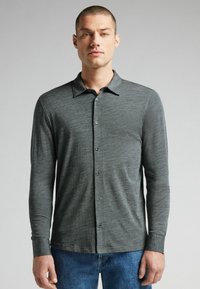 Langemouwen shirt van donkergrijze stof met een knoopsluiting aan de voorkant, klassieke kraag en een textuurafwerking. Gecombineerd met blauwe jeans.