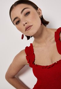 Rotes Smocked-Kleid mit Rüschenträgern, das eine taillierte Passform und eine glatte Textur aufweist. Das Modell trägt rote Ohrringe und hat einen neutralen Hintergrund.