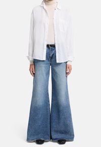 Valkoinen nappikauluspaita beigessä poolopaidassa, yhdistettynä leveälahkeisiin sinisiin farkkuihin, joissa on haalistunut gradientti ja voimakkaat ompeluyksityiskohdat.