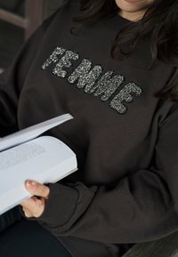 Felpa marrone con la scritta "FEMME" decorata con paillettes argento; polsini a costine, vestibilità comoda. Una persona tiene in mano un libro.