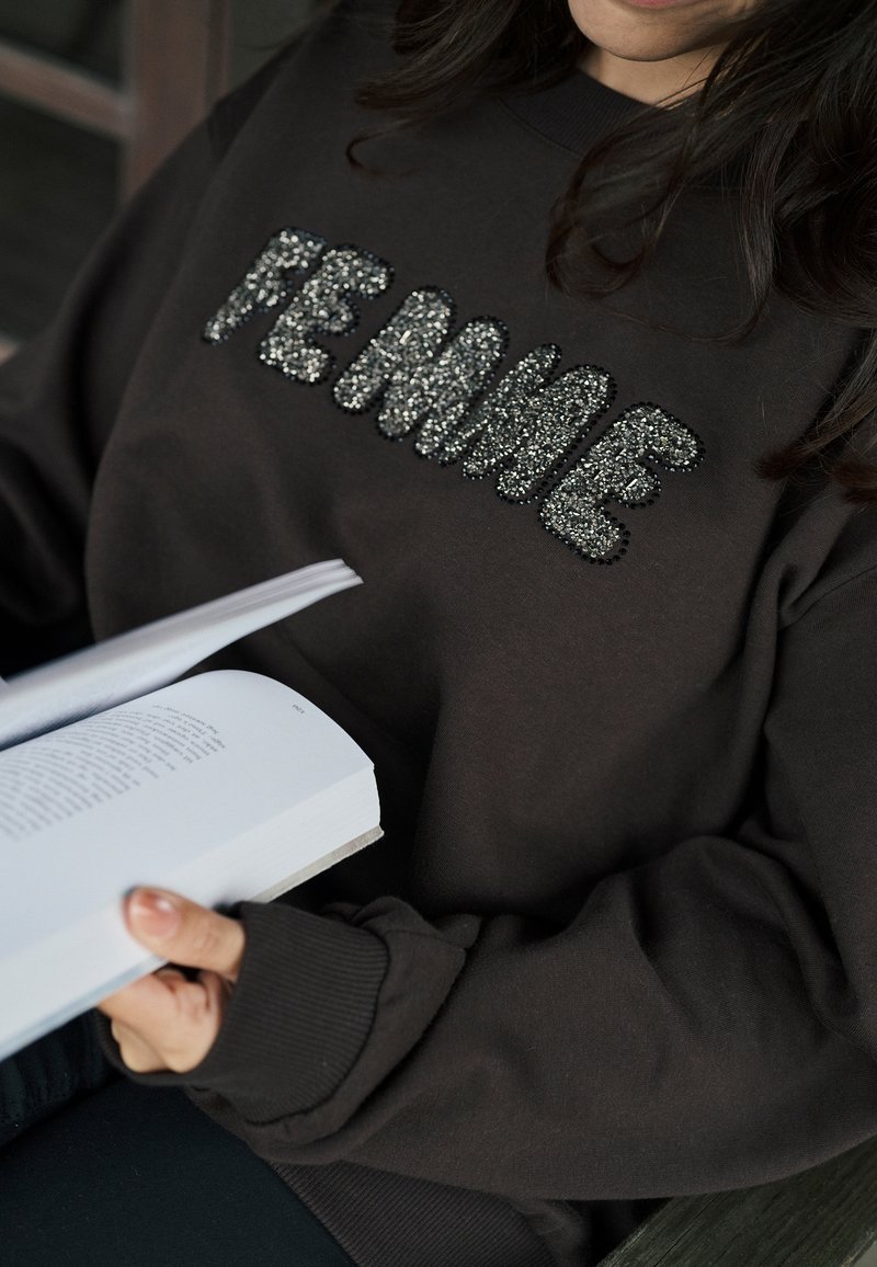 Felpa marrone con la scritta "FEMME" decorata con paillettes argento; polsini a costine, vestibilità comoda. Una persona tiene in mano un libro.