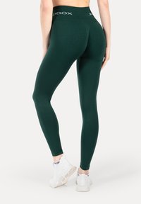 Groene leggings met een hoge tailleband met witte logotekst. Gladde textuur, nauwsluitend ontwerp en enkellange snit. Gecombineerd met witte schoenen.