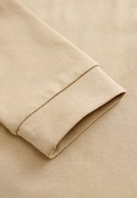 Vêtement beige à manches longues montrant un gros plan d'un poignet avec un détail en tissu côtelé et une texture lisse.