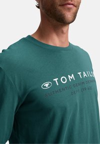 T-shirt in cotone verde acqua con colletto a giro e maniche corte, con testo frontale: "TOM TAILOR AUTHENTIC DENIM DEPARTMENT DEPT. (1962)." Texture liscia.
