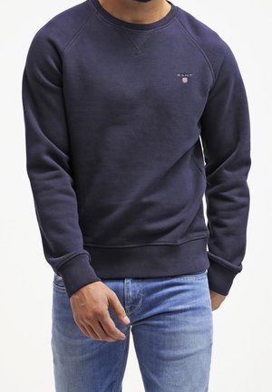 Mann trägt eine dunkelblaue GANT Sweatshirt und hellblaue Jeans mit lockerem Schnitt, steht vor einem schlichten weißen Hintergrund.