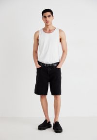 Un top alb fără mâneci, pantaloni șort negri din denim, curea neagră cu găuri și pantofi slip-on negri. Modelul stă cu mâinile în buzunare.
