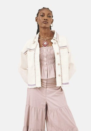 Chaqueta de mezclilla blanca con acentos bordados, combinada con una blusa sin mangas en rosa pastel y pantalones sueltos y en capas. La modelo posa con los brazos cruzados.