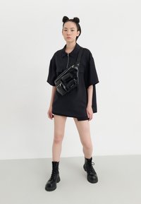 Czarna, oversize koszula z zamkiem, zestawiona z eleganckimi czarnymi butami wojskowymi i fakturowaną czarną torbą na ramię z wieloma kieszeniami.