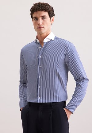 Uomo che indossa una camicia button-up a strisce verticali blu e bianche con colletto bianco, pantaloni neri e mani in tasca, in piedi davanti a uno sfondo neutro.