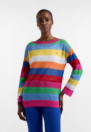 Kobieta ubrana w długi sweter dziany w wielokolorowe paski w kolorach różowym, zielonym, niebieskim, żółtym, pomarańczowym, białym i czerwonym, zestawiony z niebieskimi spodniami.