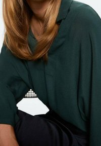 Femme aux longs cheveux châtain clair portant un blouse vert foncé et un pantalon noir, assise sur une chaise devant un fond blanc.