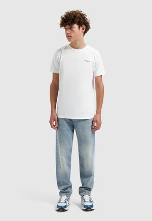 Tienerjongen met krullend haar, gekleed in een wit shirt met korte mouwen, lichtblauwe spijkerbroek en witte sneakers, staand tegen een effen achtergrond.