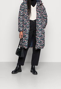 Polstret jakke med blomsterprint i marineblå, med hætte og lynlås. Kombineret med sorte korte bukser og sorte ankelstøvler, holder en lille sort taske.