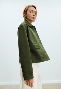 Giacca in suede verde con colletto, chiusura frontale con bottoni e tasche laterali. Design leggermente cropped con una superficie liscia.