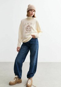 Sudadera crema con gráfico "Parque Nacional GLACIER", combinada con jeans azules holgados y botas tan con cordones. Gorro de punto beige en la cabeza.