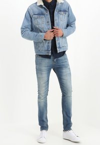 Giacca in denim azzurro chiaro con colletto in shearling, due tasche sul petto e chiusura con bottoni. Abbinata a jeans blu sbiaditi e sneakers bianche.