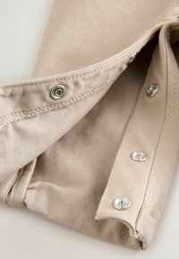 Beigenvärinen kangas, jossa hopeanväriset napsautusnapit ja saumat, esittäen vaatteen sisäpintaa, sileällä pinnalla ja siisteillä linjoilla.