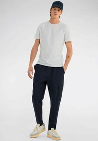 T-shirt grigio chiaro, pantaloni cargo blu marino con tasche laterali e sneakers beige con accenti verdi. Design minimale, vestibilità casual, textures di base.