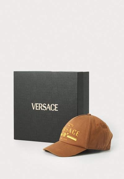 O șapcă din bumbac maro cu broderie aurie. Prezentați-o lângă o cutie texturată de culoare neagră care are numele brandului "VERSACE" scris cu aur.