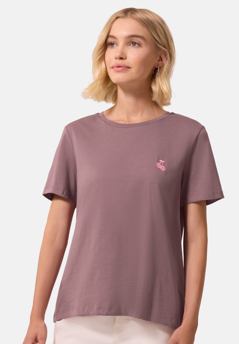 Femme aux cheveux blonds mi-longs portant un t-shirt mauve avec un petit logo cerise rose sur le côté gauche de la poitrine, debout devant un fond uni.