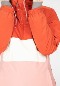 Corta vientos en bloques de colores naranja, blanco y rosa. Cuenta con un cierre de un cuarto, puños elásticos y una textura suave. Los acentos incluyen un cordón ajustable.