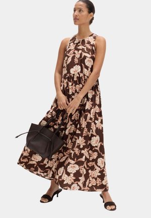 Kvinde iført en ærmeløs brun blomster maxi kjole, holder en sort håndtaske og har sorte åbne hæle på.