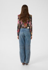 Frau mit langen braunen Haaren, die ein mehrfarbiges, floral gemustertes Langarmoberteil, hochtaillierte blaue Jeans mit gestickten Kniepaneelen und braune High Heels trägt.