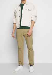 Chaqueta de mezclilla beige claro sobre una camiseta verde, combinada con pantalones caqui y zapatillas blancas. La chaqueta cuenta con dos bolsillos frontales.