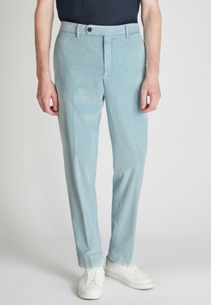 Pantalons en velours côtelé bleu clair avec des rayures verticales, fermeture éclair et bouton, présentant une coupe décontractée et une texture subtile. Baskets blanches portées.