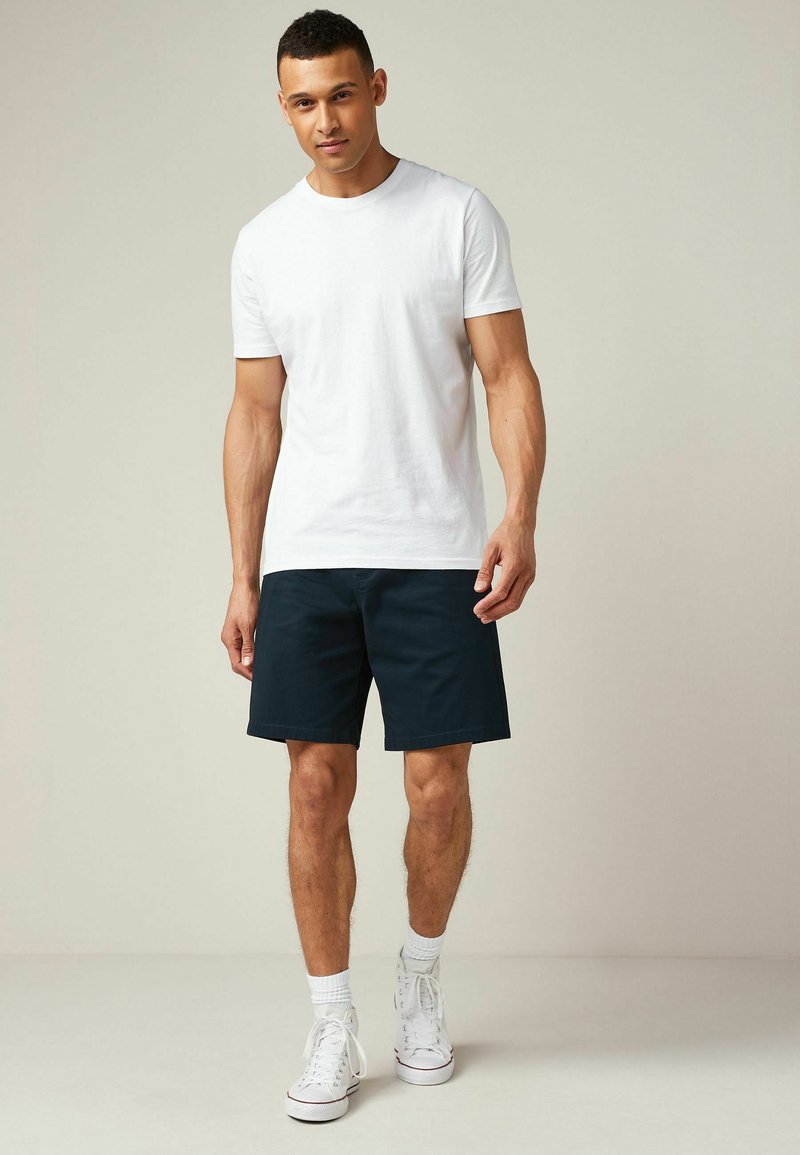 Camiseta de algodón blanca combinada con pantalones cortos azul marino, con un corte recto. El modelo lleva zapatillas blancas y calcetines hasta el tobillo.
