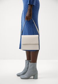 Clutch rettangolare bianco con texture a coccodrillo, tenuto in mano. Abbinato a un vestito blu a coste e a stivaletti celesti.