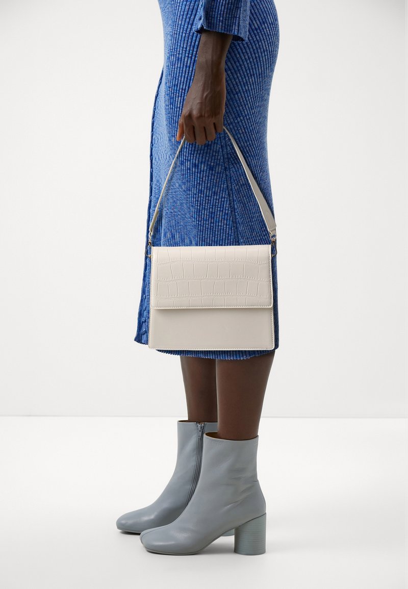 Clutch rettangolare bianco con texture a coccodrillo, tenuto in mano. Abbinato a un vestito blu a coste e a stivaletti celesti.