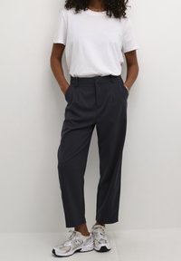 Pantalon bleu marine taille haute avec plis, associé à un t-shirt blanc uni et des baskets avec des accents argentés, offrant une coupe décontractée.