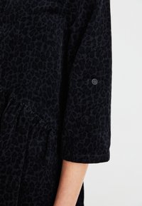 Rochie neagră din catifea cu un model de imprimeu leopar, mâneci trei sferturi și tiv cu volane, având un detaliu cu nasture pe mână.
