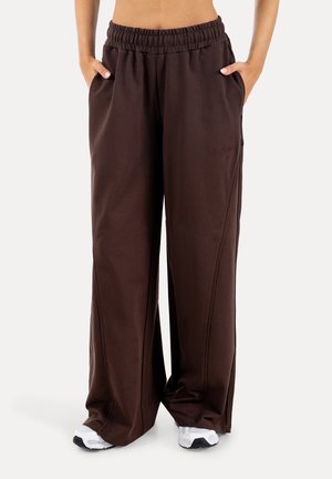 Person iført mørkebrune bukser med vidde og elastik i taljen samt hvide sneakers, hænderne i lommerne, stående på hvid baggrund.