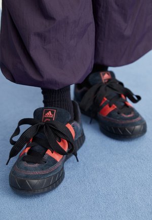 Osoba w czarno-czerwonych butach sportowych Adidas, czarnych skarpetkach i luźnych fioletowych spodniach na jasnoniebieskiej powierzchni.