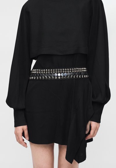 Personne portant une robe noire à manches longues et une large ceinture cloutée avec des clous argentés ronds et en forme de cône.