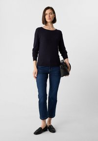 Dunkelblauer Strickpullover mit Rundhalsausschnitt, schmal geschnittene Jeans und schwarze Loafers; hält eine schwarze Umhängetasche.