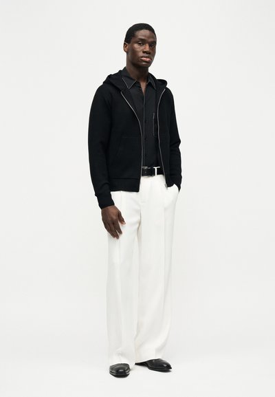 Άνδρας που στέκεται φορώντας μαύρο φούτερ με φερμουάρ, μαύρο πουκάμισο με γιακά, λευκό παντελόνι με φαρδιά ποδία, μαύρη ζώνη και μαύρα παπούτσια, μπροστά από απλό φόντο.