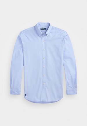 Lichtblauwe langarm blouse met knoopsluiting, voorzien van een geknoopte kraag en een donker geborduurd logo linksonder op de voorkant.