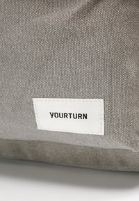Sac en tissu gris avec une surface texturée, arborant une étiquette blanche cousue avec le texte "YOURTURN" en noir. Design simple, forme rectangulaire.