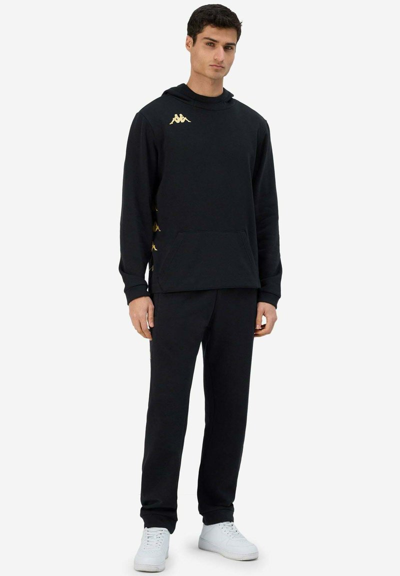 Completo felpa e pantaloni neri realizzato in tessuto morbido. La felpa con cappuccio presenta un logo dorato; i pantaloni hanno dettagli dorati abbinati sui lati. Scarpe bianche.