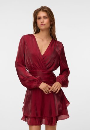 Mujer vistiendo un vestido rojo oscuro, de manga larga, con escote en V, falda con capas y cintura fruncida, de pie frente a un fondo liso.