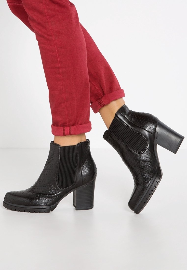 Personne portant des bottines en cuir noir texturé avec talons carrés et un pantalon rouge retroussé, marchant sur un sol clair.