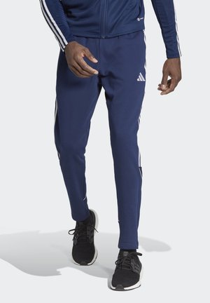 Navy blue athletic pants with white side stripes, elastic waistband, tapered legs, and a smooth texture. Black sneakers complete the look.